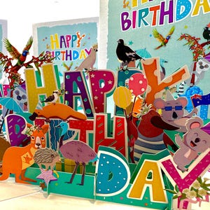 May include: A colorful pop-up birthday card featuring Australian animals and birds. The card spells out "HAPPY BIRTHDAY" with vibrant letters and includes illustrations of a kangaroo, koala, emu, and other native creatures.
