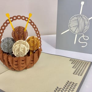 May include: A 3D pop-up greeting card featuring a brown basket filled with yarn balls in shades of brown, gray, and yellow. Yellow knitting needles are placed in the yarn balls. A gray card with a yarn ball and needles design is in the background.