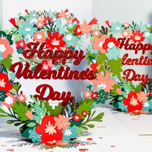 May include: A red glitter paper Valentine's Day card with a heart-shaped wreath of flowers and leaves. The card reads "Happy Valentine's Day".