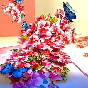 Puede incluir: Una tarjeta de felicitación emergente con una vibrante exhibición de orquídeas rojas y blancas y mariposas azules. La tarjeta está abierta, revelando un arreglo floral tridimensional. Se ven tarjetas adicionales con diseños similares.