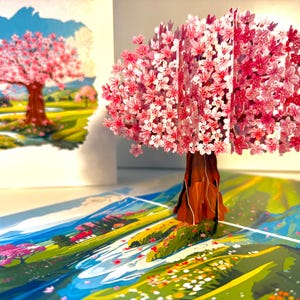 May include: A 3D pop-up card featuring a cherry blossom tree with pink and white flowers. The tree is brown with a detailed trunk and branches. The card is open, revealing a colorful landscape with a river and other trees.