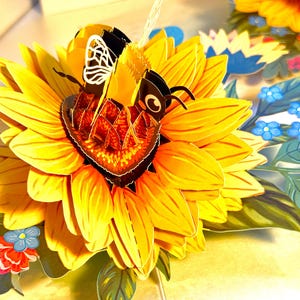 May include: A 3D pop-up card featuring a vibrant yellow sunflower with a detailed bee. The bee has black and yellow stripes and is perched on the flower's center. Additional flowers and leaves in blue, red, and green surround the sunflower.