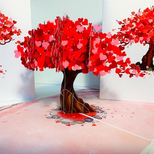 May include: A 3D pop-up card featuring a red heart-shaped tree with a brown trunk and a pink background. The tree is surrounded by small red hearts.