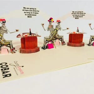 May include: A pop-up greeting card featuring skeleton figures seated at tables, holding drinks. The card has the text "We are going to sit here and drink for as long as it takes to understand MEN!" and the word "COBAR".