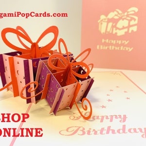 Puede incluir: Una tarjeta de cumpleaños 3D pop-up con una pila de cajas de regalo con lazos rojos y detalles en rosa y morado. La tarjeta está abierta y las cajas están saliendo. La tarjeta también tiene el texto "Feliz cumpleaños" en el frente.