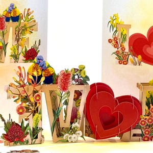 Puede incluir: Una tarjeta de felicitación desplegable con la palabra "LOVE" en letras de madera, decorada con flores coloridas, pájaros y formas de corazón rojo. La tarjeta está diseñada con un efecto 3D, creando un diseño visualmente atractivo y artístico.