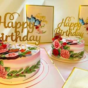 May include: A pink and white cake with a gold glitter "Happy Birthday" topper and a blue butterfly. The cake is decorated with pink and red roses and green leaves. The cake is on a white plate with a pink border.
