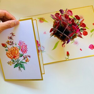 May include: A pop-up card with a 3D bouquet of red, pink, and purple roses. The card is open and the front panel shows a flat illustration of a bouquet of roses in shades of orange, pink, and green.
