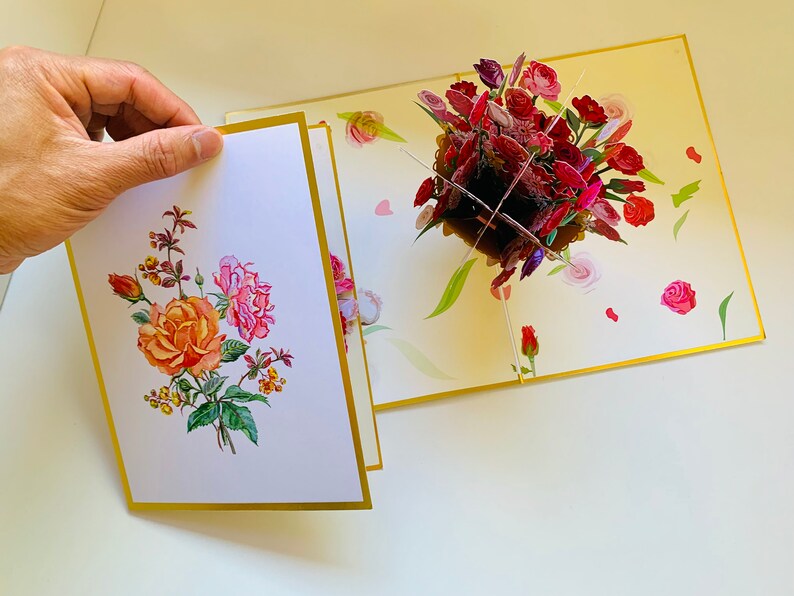May include: A pop-up card with a 3D bouquet of red, pink, and purple roses. The card is open and the front panel shows a flat illustration of a bouquet of roses in shades of orange, pink, and green.