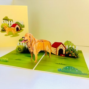 May include: A 3D pop-up greeting card featuring a golden retriever dog. The card depicts a doghouse, a white picket fence, and green grass. The dog is light brown with a pink tongue. The card is open and standing upright.