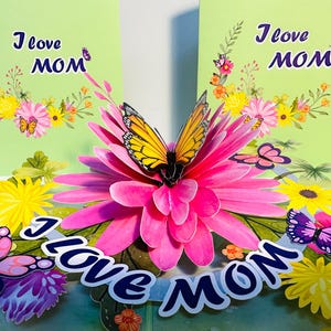 May include: A 3D pop-up card with a pink flower and butterflies. The card is decorated with green leaves and yellow flowers. The card reads "I love Mom" in blue letters.