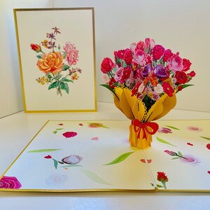 May include: A 3D pop-up card featuring a bouquet of pink, red, and purple flowers in a yellow vase with a red bow. The card is open and the flowers are popping up from the vase. The card has a white background with a gold border.