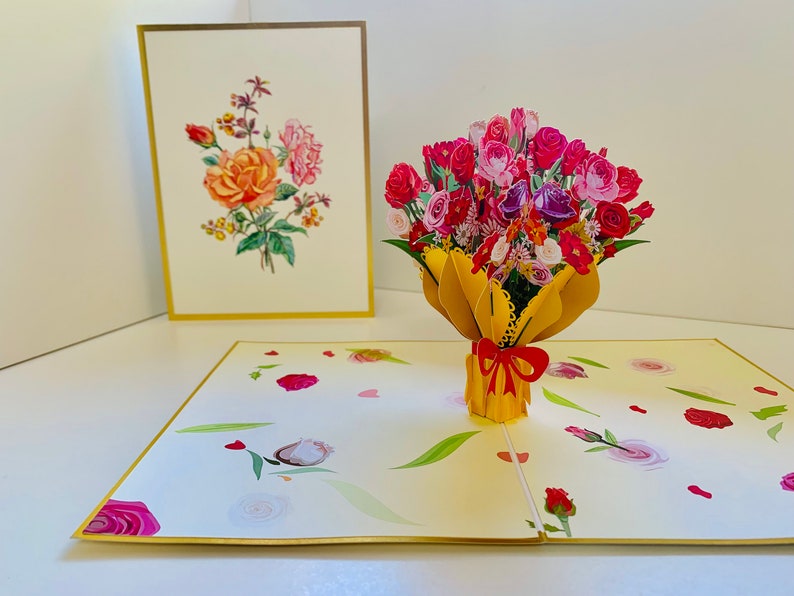 May include: A 3D pop-up card featuring a bouquet of pink, red, and purple flowers in a yellow vase with a red bow. The card is open and the flowers are popping up from the vase. The card has a white background with a gold border.