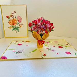 May include: A 3D pop-up card featuring a bouquet of pink, red, and white roses in a yellow vase. The card is open and the roses are in focus. The background of the card is a light yellow with a pattern of small pink and white flowers.