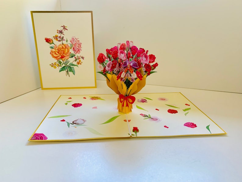 May include: A 3D pop-up card featuring a bouquet of pink, red, and white roses in a yellow vase. The card is open and the roses are in focus. The background of the card is a light yellow with a pattern of small pink and white flowers.