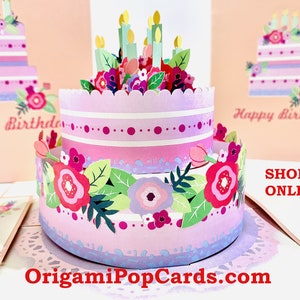 Puede incluir: Una tarta de cumpleaños de papel rosa de dos pisos con decoraciones florales y velas verdes. La tarta está rodeada de tarjetas a juego que dicen "Feliz Cumpleaños". El sitio web OrigamiPopCards.com es visible.
