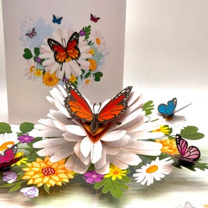Puede incluir: Tarjeta de felicitación emergente 3D con una gran margarita blanca y una mariposa naranja y negra. Mariposas adicionales en azul, rosa y rojo están dispersas alrededor del arreglo floral. La tarjeta tiene un tema floral.