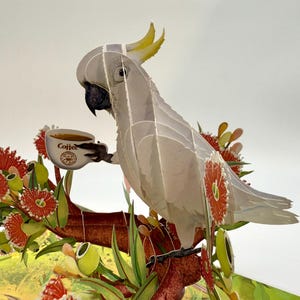 May include: A 3D paper art piece featuring a white cockatoo holding a coffee cup. The bird is perched on a branch with red and green foliage. The coffee cup has the word "Coffee" printed on it.