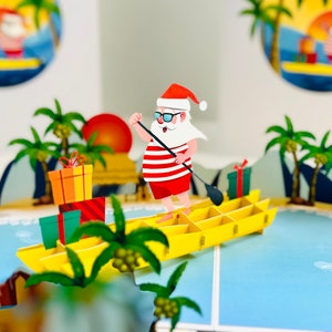 Puede incluir: Tarjeta de felicitación desplegable con Papá Noel haciendo paddleboard en una tabla amarilla. Papá Noel lleva una camisa a rayas rojas y blancas, pantalones cortos rojos y un gorro de Papá Noel. Escena de playa tropical.