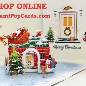 May include: A 3D pop-up Christmas card featuring a red brick house with a green roof, a snowman, a dog, a Christmas tree, and Santa Claus in a sleigh pulled by reindeer. The card is open and the pop-up elements are visible. The text "Merry Christmas" is printed on the card. The text "SHOP ONLINE OrigamiPopCards.com" is printed at the top of the card.