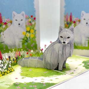 Puede incluir: Una tarjeta emergente 3D con una figura de zorro gris sentada en un área de césped verde con flores coloridas. El fondo muestra una pintura de un zorro blanco en un jardín de flores bajo un cielo azul. La tarjeta está abierta y expuesta sobre una superficie blanca.