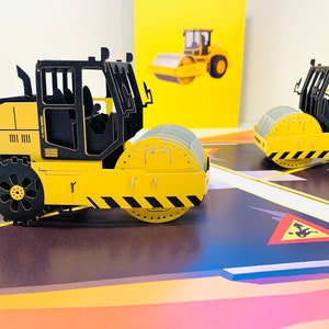 May include: A yellow and black construction vehicle with a large roller in the front. The vehicle is a paper model and is part of a pop-up card.