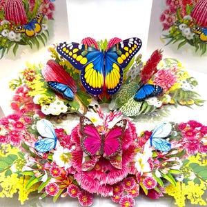 Puede incluir: Una tarjeta de felicitación desplegable con un vibrante arreglo floral y mariposas de colores. La tarjeta presenta una exhibición central de flores rosas, rojas, amarillas y blancas, con varias mariposas azules y rosas. Las mariposas tienen intrincados patrones de alas.