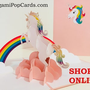 Puede incluir: Una tarjeta pop-up 3D con un unicornio blanco saltando sobre un arcoíris con nubes rosas. La tarjeta está abierta y el unicornio está en el centro de la imagen. La tarjeta está sobre un fondo blanco. El texto "SHOP ONLINE" está en letras rojas en la parte inferior de la tarjeta.