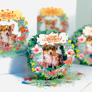 Puede incluir: Una tarjeta 3D pop-up con una corona de flores con dos corgis dentro. La tarjeta tiene el texto "Happy Mother's Day" en la corona.