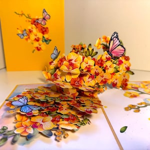 May include: A 3D pop-up greeting card with a floral design of yellow and orange orchids and butterflies. The card is open, showcasing the detailed floral arrangement, with a matching yellow background card.