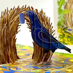 May include: A 3D paper art piece featuring a dark blue bird holding a blue bottle cap in its beak. The bird stands in front of brown paper trees, with a colorful illustrated background. The artwork is a pop-up style, with intricate details.