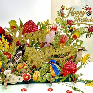 May include: A pop-up birthday card featuring colorful birds, flowers, and foliage. The words "Happy Birthday" are in gold glitter. Birds include a red parrot, a black and white magpie, a blue kingfisher, and a pink cockatoo.