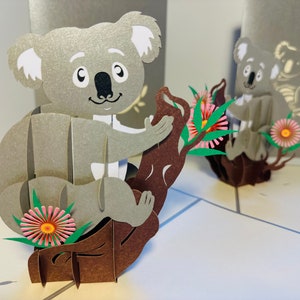 May include: 3D pop-up card showing a grey koala bear on a brown branch with pink and yellow flowers. The koala has white ears and a smiling face. Another koala is visible in the background.