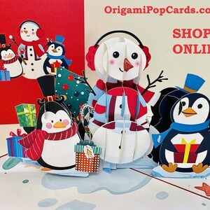 May include: A 3D pop-up Christmas card featuring a snowman and penguins. The snowman is wearing a red and blue scarf and a blue hat. The penguins are wearing scarves and hats. The card is decorated with a Christmas tree and presents. The text "OrigamiPopCards.com SHOP ONLINE" is printed on the card.