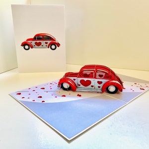 May include: A red pop-up card featuring a red Volkswagen Beetle covered in white hearts. The card is open and the car is in a 3D pop-up position. The background of the card is a light blue with a scattering of red hearts.