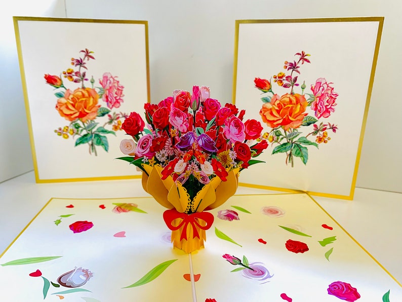 May include: A 3D pop-up card featuring a bouquet of red, pink, and white roses in a yellow paper wrapper with a red bow. The card is open and the roses are popping up from the center. The card is surrounded by smaller roses and hearts.