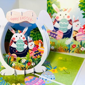 Puede incluir: Tarjeta de Pascua 3D con diseño de conejo blanco y la frase "Happy Easter!". En el interior, conejos, huevos coloridos y una escena de jardín. La tarjeta abierta revela un diseño desplegable.
