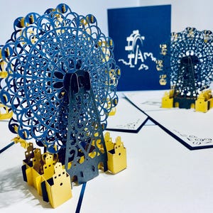 Puede incluir: Tarjeta desplegable con un diseño de noria en azul y amarillo. El intrincado arte en papel muestra la estructura y las góndolas de la rueda. Una tarjeta azul oscuro con texto blanco y un gráfico de noria está en el fondo.