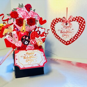 May include: A red and pink pop-up card with a bouquet of roses, chocolates, champagne, and a gift box. The card says "Happy Valentine's Day" on the front and inside. The card is decorated with hearts and polka dots.