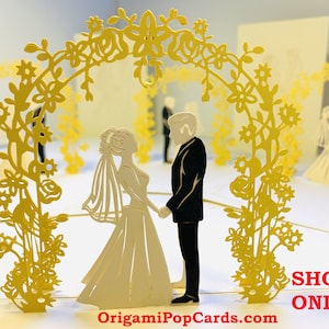 May include: A 3D pop-up card depicting a wedding ceremony with a bride and groom standing beneath a floral archway. The card is crafted from white and yellow paper and features a delicate, intricate design. The text "OrigamiPopCards.com" and "SHOP ONLINE" is printed on the card.