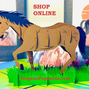 Puede incluir: Tarjeta desplegable de papel marrón con un caballo con crin y cola azules, sobre un paisaje con hierba verde y montañas. El texto "SHOP ONLINE" está en rojo en la parte superior.