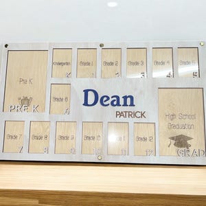 May include: A wooden school years display board with spaces for photos from Pre-K to High School Graduation. The name "Dean Patrick" is displayed in blue letters. The board is light-colored with a wooden frame.