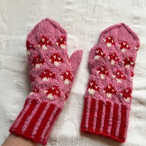 May include: A pair of pink knitted mittens with a red and white floral pattern. The cuffs are red with vertical stripes. The mittens are made of a soft, warm material, perfect for cold weather.