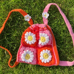 May include: A handmade crochet backpack featuring a daisy pattern in shades of orange, pink, and white. The bag has orange and pink straps, and is decorated with small white flower accents. The bag is sitting on a bed of green grass.
