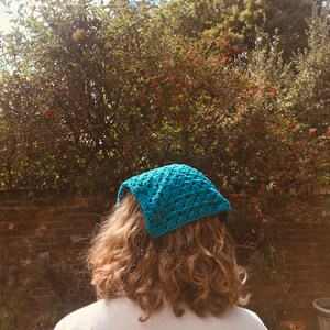 May include: A person with curly hair wearing a teal blue crocheted headband. The headband is tied at the back of the head and covers the forehead.