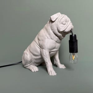 Puede incluir: Una escultura de perro pug de cerámica blanca con un cable negro y una bombilla en la boca. La escultura está sentada sobre una superficie verde.