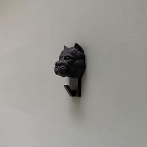 May include: A black metal wall hook in the shape of a dog's head. The hook is a dark, matte black and features a detailed sculpture of a dog's face, with a hook extending downwards. The hook is mounted on a light-coloured wall.