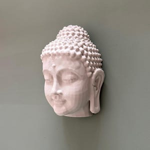 May include: A white Buddha head sculpture with closed eyes and a serene expression. The sculpture has detailed features, including a textured hairstyle and delicate facial contours. The head is mounted on a muted green wall, creating a calming aesthetic.