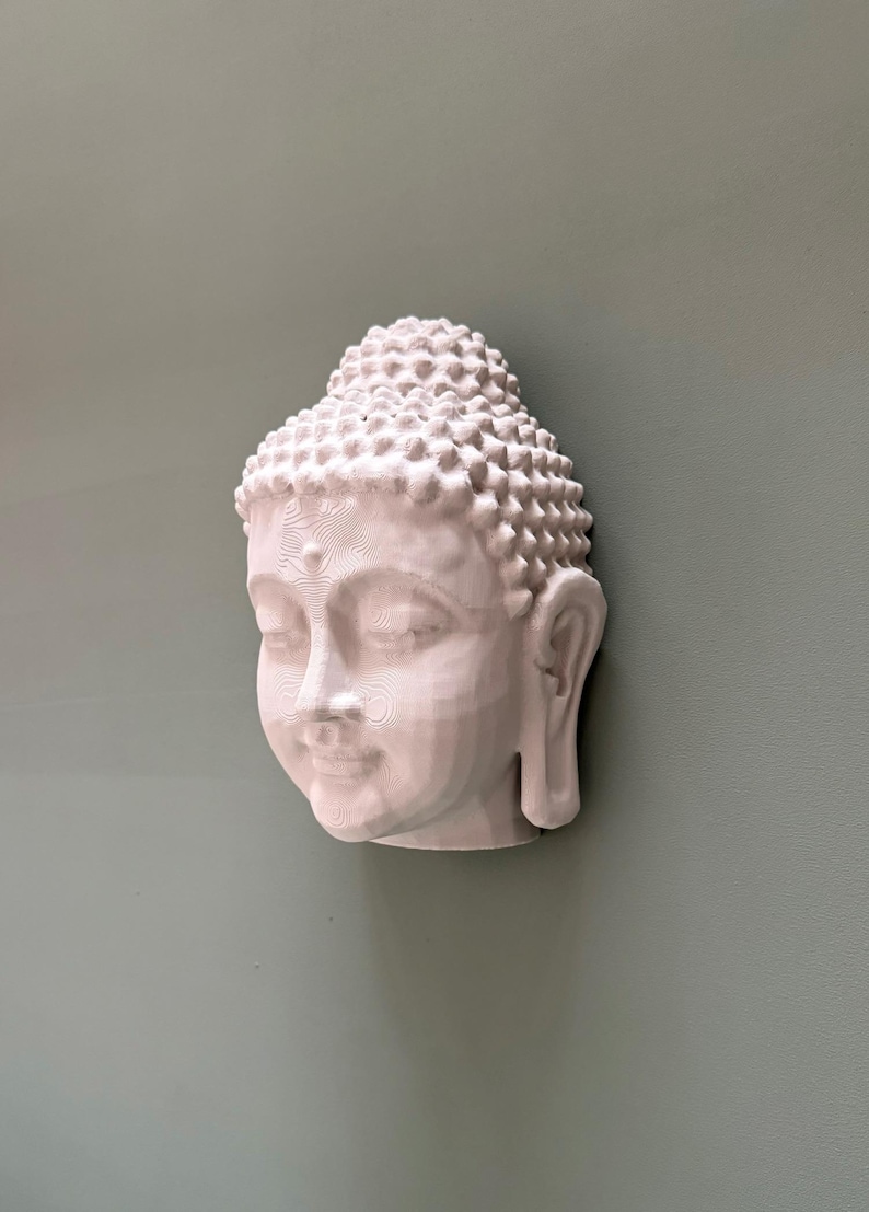 May include: A white Buddha head sculpture with closed eyes and a serene expression. The sculpture has detailed features, including a textured hairstyle and delicate facial contours. The head is mounted on a muted green wall, creating a calming aesthetic.