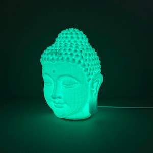 以下が含まれることがあります： 緑色に光る仏陀の頭部彫刻。彫刻は、目を閉じた穏やかな表情とテクスチャ加工された表面が特徴です。光は内側から発せられ、柔らかな輝きを生み出します。ベースからワイヤーが伸びています。